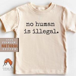 May include: A beige short-sleeved t-shirt with the text "no human is illegal." printed in black. The shirt is made of 100% premium natural cotton.