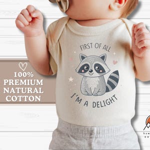 May include: A white baby bodysuit with a cute raccoon graphic and the text "First of all, I'm a delight". The bodysuit is made of 100% premium natural cotton.