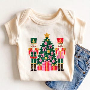 May include: A cream-colored ribbed baby bodysuit featuring a central graphic of a green Christmas tree decorated with pink bows and gold ornaments, flanked by two nutcracker figures in red and green uniforms with gift boxes.