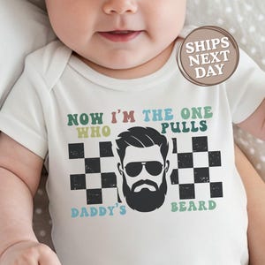 May include: White baby bodysuit with the text "NOW I'M THE ONE WHO PULLS DADDY'S BEARD" in green, blue, and black. The design includes a graphic of a bearded man with sunglasses and a checkered pattern.
