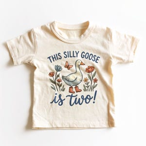May include: A cream-colored short-sleeve t-shirt featuring a graphic of a white goose wearing orange boots, surrounded by colorful flowers and a butterfly. The text reads "THIS SILLY GOOSE is two!" in dark blue lettering centered on the front.