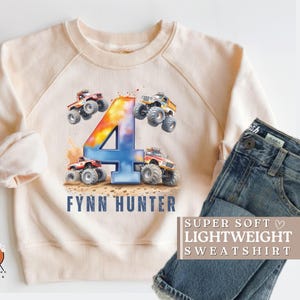 May include: A light beige sweatshirt with a colorful number four and monster truck graphic. The text "Fynn Hunter" is printed below the graphic. The text "SUPER SOFT LIGHTWEIGHT SWEATSHIRT" is printed on a separate piece of fabric.