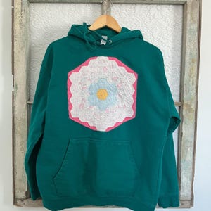 May include: A teal hoodie with a large, colorful floral patch on the front. The patch features a white flower with blue and yellow accents, framed by pink and white geometric shapes. The hoodie has a kangaroo pocket and a drawstring hood.
