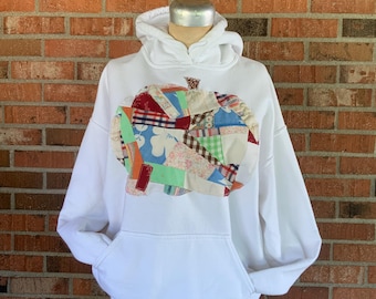 May include: White hoodie featuring a patchwork pumpkin design in various colours. The pumpkin is made from fabric scraps in red, blue, green, and orange. The sweatshirt has a kangaroo pocket and a hood.