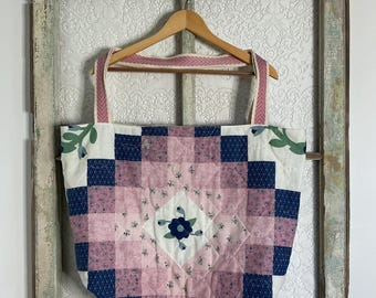 May include: A quilted tote bag featuring a patchwork design in shades of pink, navy blue, and white. The bag displays a central floral motif, a geometric block pattern, and two wide fabric straps with a pink and white chevron-style weave pattern.