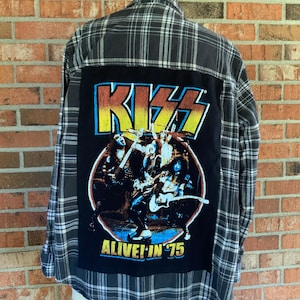 May include: A gray and black plaid flannel shirt with a large black patch featuring the band KISS. The patch has the band's logo in yellow, blue, and red, with the words "ALIVE! IN '75". The shirt is displayed on a mannequin.
