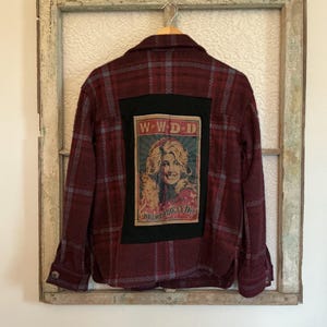 May include: A maroon plaid jacket with a black square patch featuring a portrait of Dolly Parton and the text "WWDD". The jacket is hanging on a wooden hanger against a vintage window frame.