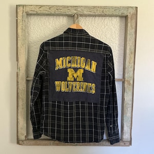May include: A long-sleeve button-down shirt in a black and grey plaid pattern. The back features a large, rectangular dark grey fabric patch with the yellow text "MICHIGAN" and "WOLVERINES" along with a yellow "M" logo in the center.