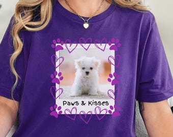 May include: A purple t-shirt featuring a framed image of a white puppy with the text "Paws & Kisses". The frame is decorated with purple hearts and paw prints. The shirt is a solid purple color.