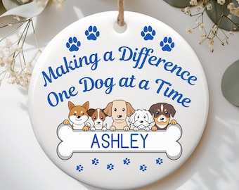 May include: A round white ceramic ornament hanging from a jute string. The text "Making a Difference One Dog at a Time" is printed in blue above five illustrated dogs standing behind a large white bone with the name "ASHLEY" in blue text.