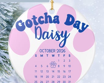 May include: A round white ceramic ornament featuring a large light pink paw print design. The text reads Gotcha Day Daisy and October 2026 with a calendar grid. A small paw print icon marks the 7th day. A gold string is attached at the top.