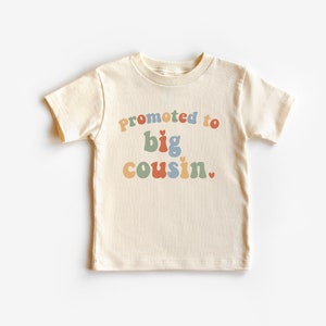 May include: Cream-colored toddler t-shirt with the phrase "Promoted to Big Cousin" in retro-style, multi-colored lettering. The design includes a small heart at the end of the text. The shirt is made of soft material.