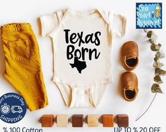May include: A white baby onesie with black text that reads "Texas Born" and a black outline of the state of Texas. The onesie is on a white background with brown leather baby shoes, wooden blocks, and a sprig of greenery.