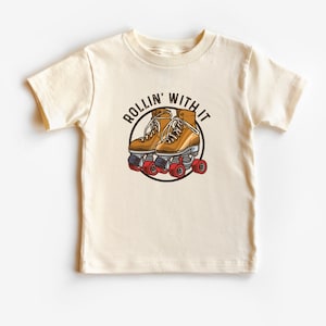 May include: A cream colored t-shirt with a graphic of a pair of roller skates and the text "Rollin' With It".