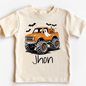 May include: Cream-colored t-shirt featuring an orange monster truck with a ghost and pumpkin graphic. The truck has large black tires and is decorated with bats. The name "Jhon" is printed in black script below the truck.