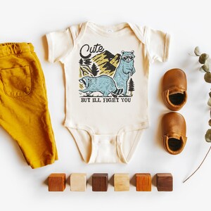 Pu&ograve; includere: Un body bianco per neonato con una grafica di un procione e un castoro in un ambiente boschivo. Il testo sul body recita "Cute but I'll fight you". Il body &egrave; abbinato a pantaloni color giallo senape e scarpine da bambino in pelle marrone.