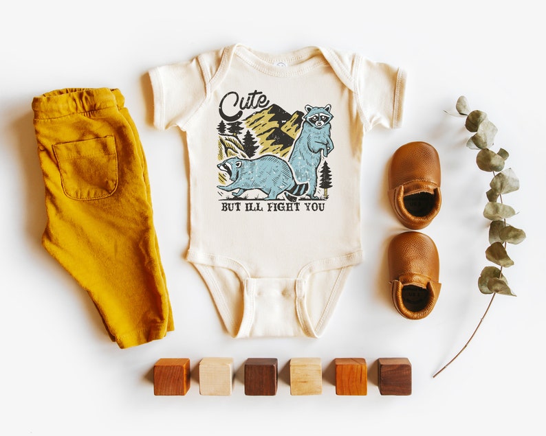 Pu&ograve; includere: Un body bianco per neonato con una grafica di un procione e un castoro in un ambiente boschivo. Il testo sul body recita "Cute but I'll fight you". Il body &egrave; abbinato a pantaloni color giallo senape e scarpine da bambino in pelle marrone.