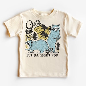 Pu&ograve; includere: Una maglietta beige con una grafica di due procioni in un ambiente boschivo. Il testo "Cute but I'll fight you" &egrave; stampato sotto i procioni.