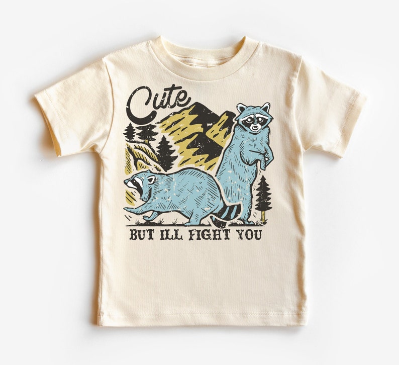 Pu&ograve; includere: Una maglietta beige con una grafica di due procioni in un ambiente boschivo. Il testo "Cute but I'll fight you" &egrave; stampato sotto i procioni.