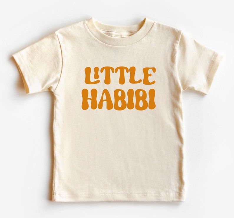 May include: A cream coloured short-sleeved t-shirt with the text "LITTLE HABIBI" in orange.