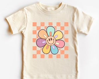 May include: A cream colored t-shirt with a rainbow colored flower with a smiley face in the center. The flower is on a checkered background with orange and white squares.