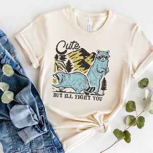 Pu&ograve; includere: Una maglietta beige con una grafica di due procioni in un ambiente boschivo. Il testo "Cute but I'll fight you" &egrave; stampato sotto i procioni.