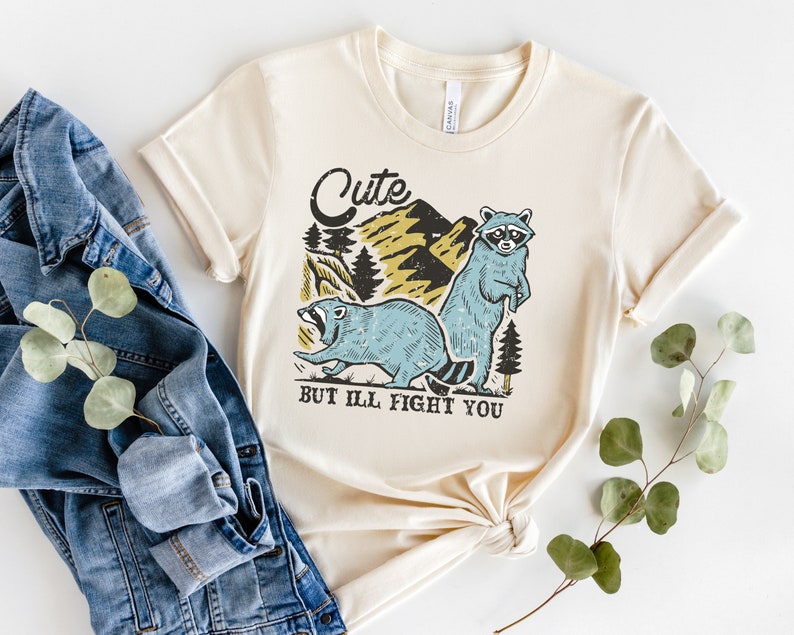 Pu&ograve; includere: Una maglietta beige con una grafica di due procioni in un ambiente boschivo. Il testo "Cute but I'll fight you" &egrave; stampato sotto i procioni.