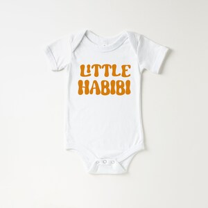 May include: White baby onesie with short sleeves and a brown text graphic that says "LITTLE HABIBI".