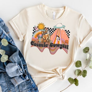 以下が含まれることがあります： クリーム色のTシャツに、カウガールが馬に乗っているレトロなグラフィックデザインが施されています。虹、UFO、そして「Cosmic Cowgirl」の文字が描かれています。