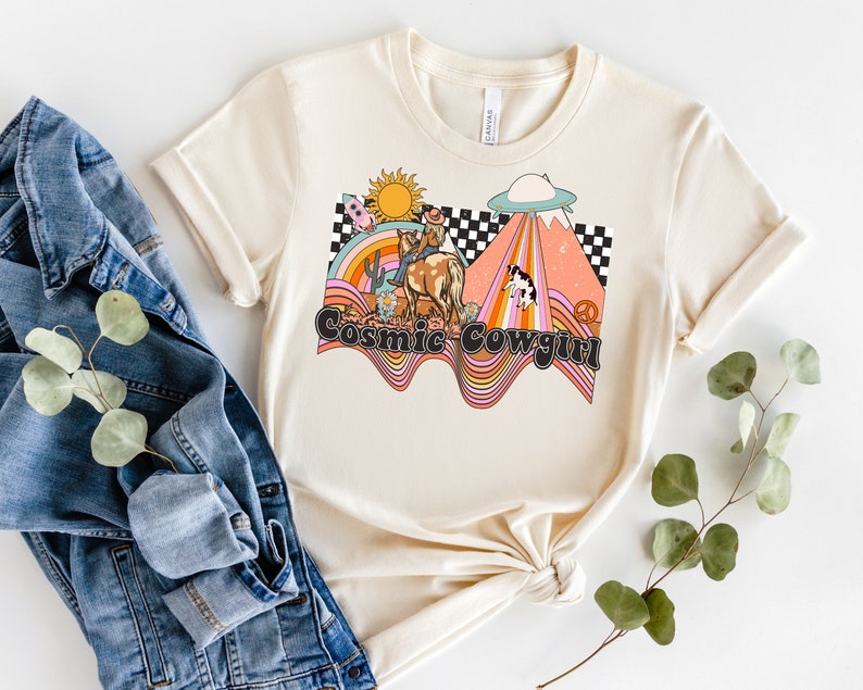 以下が含まれることがあります： クリーム色のTシャツに、カウガールが馬に乗っているレトロなグラフィックデザインが施されています。虹、UFO、そして「Cosmic Cowgirl」の文字が描かれています。