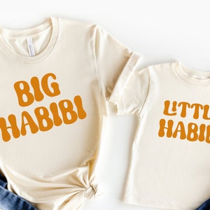 May include: Two cream coloured t-shirts with the text "BIG HABIBI" and "LITTLE HABIBI" in orange. The shirts are laid out on a white background with blue jeans and dried flowers.