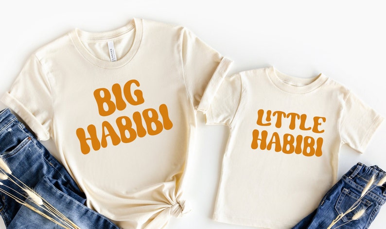 May include: Two cream coloured t-shirts with the text "BIG HABIBI" and "LITTLE HABIBI" in orange. The shirts are laid out on a white background with blue jeans and dried flowers.