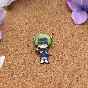 May include: A colorful enamel pin featuring a cartoon character wearing a black shirt, blue pants, and white shoes, with a green alien head. The pin is set on a brown corkboard, surrounded by purple flowers and green leaves.