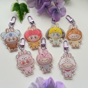 May include: Acrylic keychains featuring cartoon characters in various animal costumes, including a koala, apple, and bunny. Each keychain has a silver-colored clasp. The keychains are in shades of brown, pink, yellow, and orange.