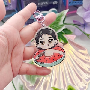 May include: A clear acrylic keychain featuring a cartoon character with black hair, wearing a watermelon-shaped inflatable pool float. The character has a light pink blush and is smiling.