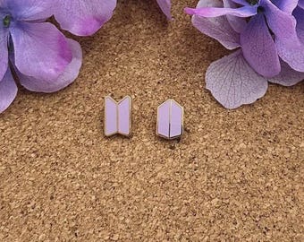 Puede incluir: Un par de pendientes de botón con acabado en esmalte de color violeta claro y borde metálico. Uno tiene forma de logotipo trapezoidal y el otro es pentagonal, ambos con un detalle de línea vertical en el centro.