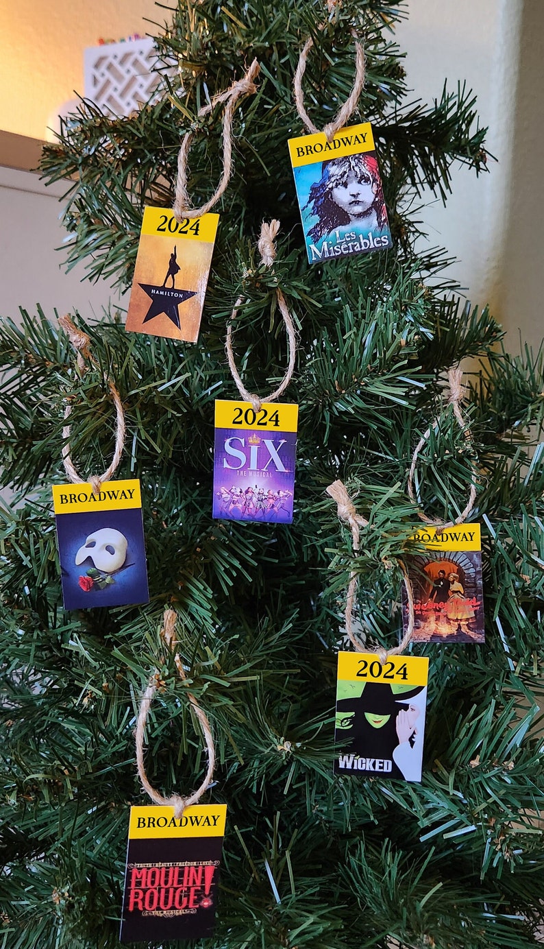May include: A Christmas tree decorated with six ornaments featuring Broadway show posters. The ornaments are yellow with black text and images. The shows featured are Hamilton, Les Miserables, Six, Phantom of the Opera, Wicked, and Moulin Rouge.