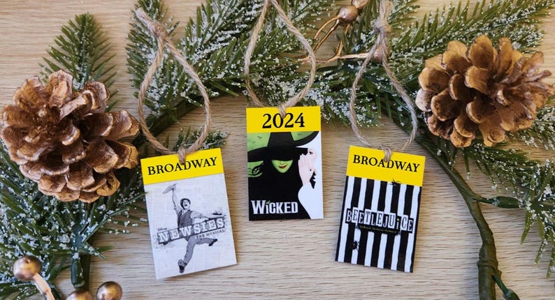 May include: Three Christmas ornaments with yellow and black Broadway show themes. The ornaments feature the text "Broadway" and the names of the shows "Newsies", "Wicked", and "Beetlejuice". The ornaments are decorated with images from the shows and the year 2024.