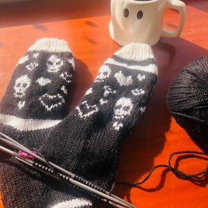 May include: A pair of black and white knit socks with a skull and heart pattern. The socks are being knitted with two knitting needles and a ball of black yarn is in the background.