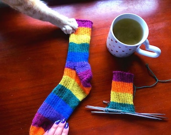 May include: A completed rainbow-striped sock next to a partially knitted sock. The finished sock is held by a hand with purple nail polish and a cat's paw. A mug of tea with heart-shaped details sits nearby.