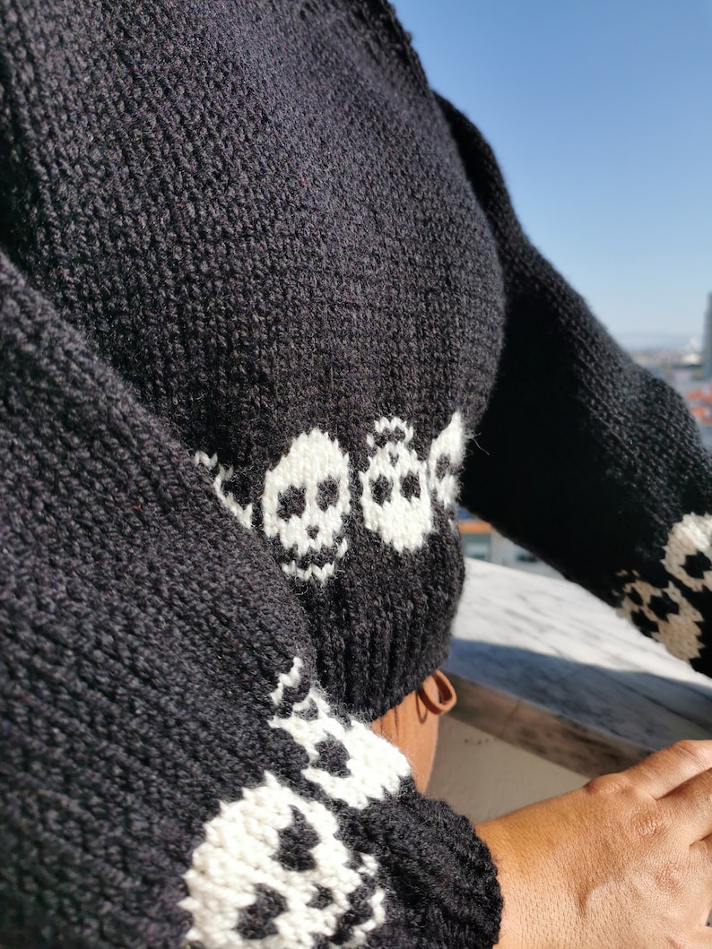K&ouml;nnte beinhalten: Schwarzer Strickpullover mit wei&szlig;en Totenkopf-Designs. Der Pullover hat lange &Auml;rmel und eine verk&uuml;rzte L&auml;nge.