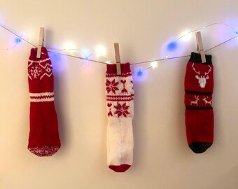 Peut inclure: Trois chaussettes de Noël rouges et blanches avec des motifs festifs, suspendues à une guirlande lumineuse. Chaque chaussette est fixée avec une pince à linge en bois. Motifs de flocons de neige et de rennes.