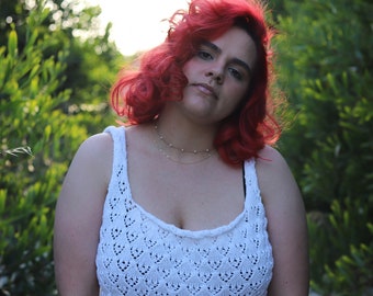 Könnte beinhalten: Ein weißes Häkeltop mit offenem Stich und rundem Ausschnitt. Das Oberteil wird mit grauen Jeans kombiniert. Die Person auf dem Bild hat leuchtend rotes Haar und trägt eine zarte goldene Halskette. Der Hintergrund ist mit grünem Laub verschwommen.