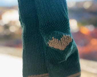 Könnte beinhalten: Ein Paar handgestrickte Socken in Türkisgrün mit beigen Akzenten. Die Socken haben einen beigen Streifen an der Ober- und Unterseite sowie ein herzförmiges Design in Beige an der Ferse. Das Garn ist weich und strukturiert.