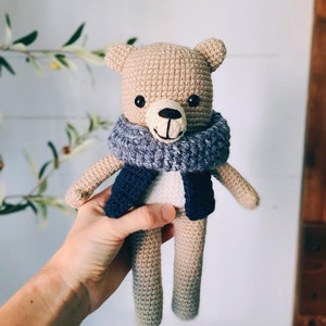 May include: A crocheted teddy bear wearing a blue and gray scarf and a blue vest. The bear has black eyes and a black nose.