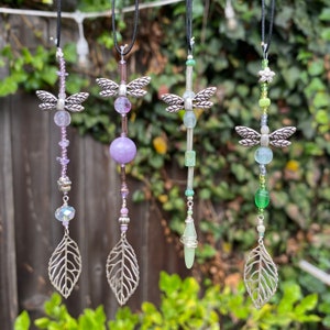 May include: Five silver dragonfly suncatchers with various colored beads and crystals. The suncatchers are hanging from black cords against a green leafy background.