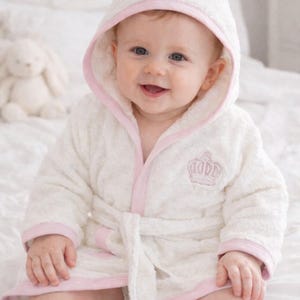 May include: A smiling baby wearing a white hooded bathrobe with pink trim. The hood is embroidered with the name "EMILY" in pink. A pink crown is embroidered on the front of the robe. The baby is sitting on a white surface.