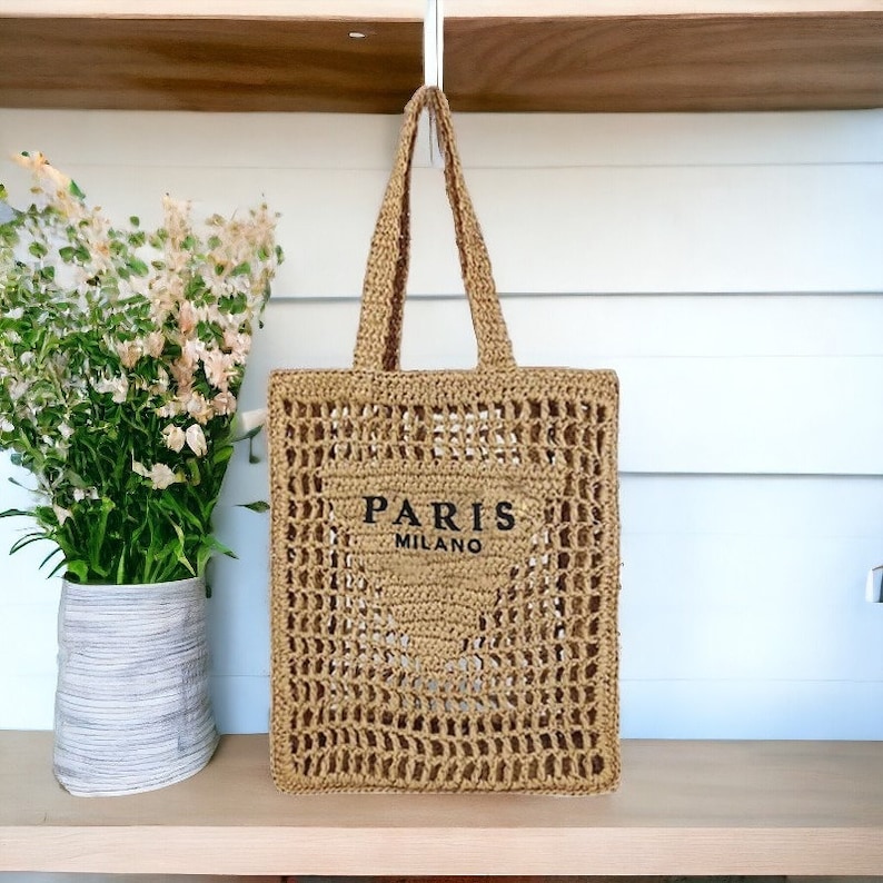 May include: A woven beige tote bag with the words "PARIS MILANO" printed in black. The bag has a rectangular shape with a woven handle and a large open weave design. The bag is sitting on a wooden shelf.