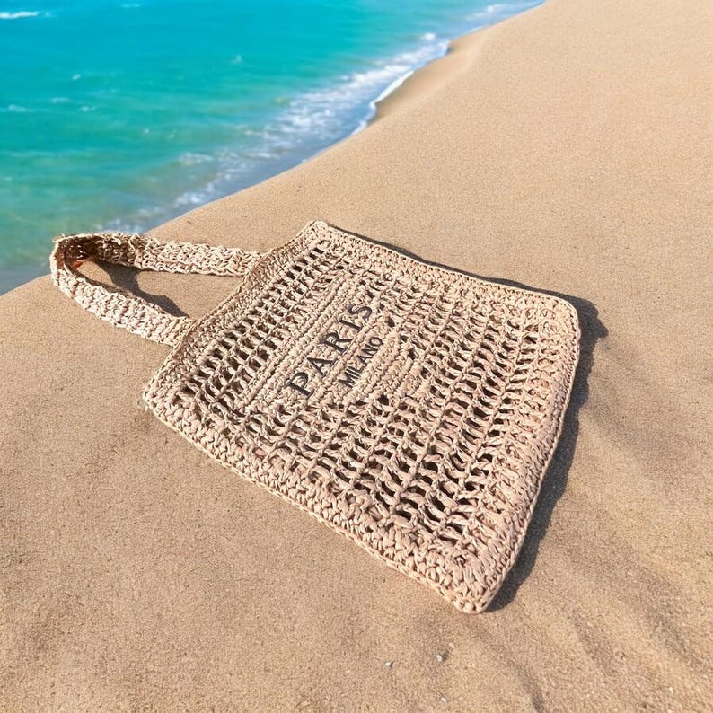 May include: A beige woven tote bag with a handle, resting on sand near the ocean. The bag features an open weave design and the text "PARIS MILANO" in black.