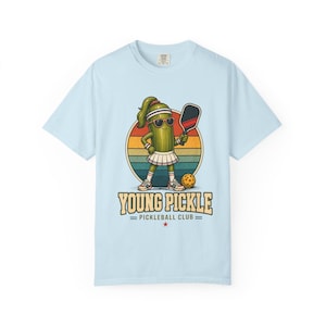 May include: Light blue t-shirt featuring a cartoon pickle character wearing sunglasses, a headband, and a tennis skirt, holding a pickleball paddle. The design includes the text "YOUNG PICKLE = PICKLEBALL CLUB =" with a retro sunset background.